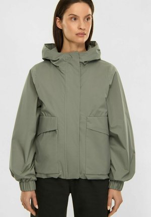 Woman wearing a light green hooded jacket with front pockets and elastic cuffs, paired with black pants, standing against a plain background.