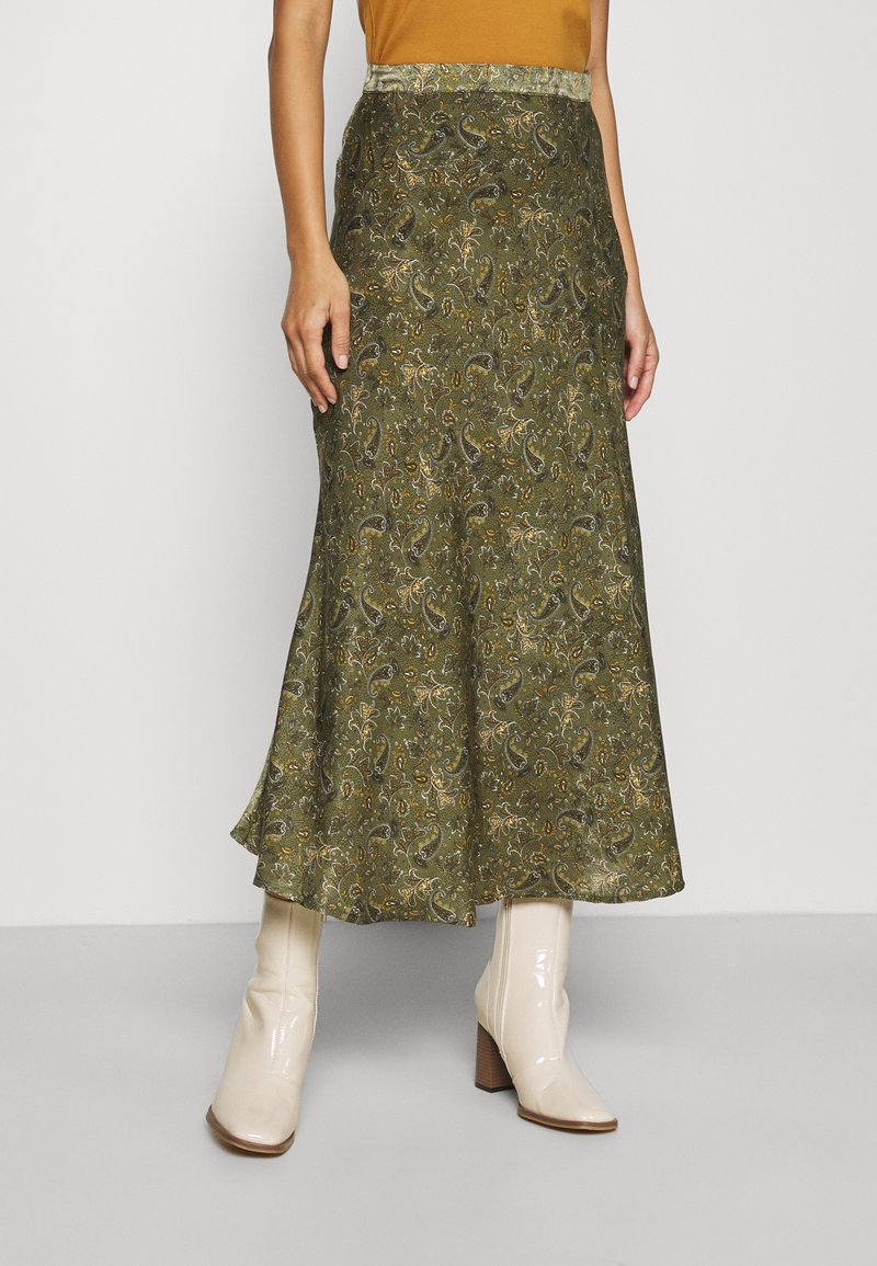 Femme portant une jupe midi verte à motif cachemire et des bottines en cuir verni crème à talons blocs sur un fond uni.