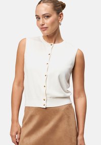 Pull blanc sans manches avec col rond, doté d'une rangée de boutons dorés sur le devant et d'un ourlet côtelé. Texture douce, design ajusté.