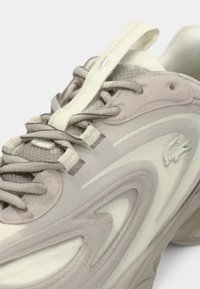 Chaussure de sport gris clair composée de mesh et de caoutchouc, avec un design incurvé, des accents texturés et un logo sur le côté.