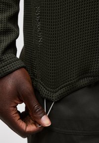 Mână cu piele închisă reglând șnurul unei jachete texturate verde măsliniu, cu logo-ul INOV-8 vizibil de-a lungul cusăturii laterale.