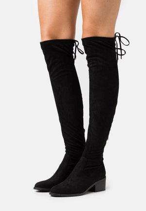 Stivali neri in suede alti fino alla coscia con tacco basso e lacci posteriori, indossati su gambe nude su uno sfondo bianco semplice.