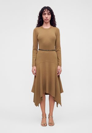 Une femme porte une robe en maille camel à manches longues et à l’ourlet asymétrique, une ceinture chaîne et des sandales dorées, sur un fond uni.