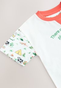 Balti marškinėliai su koralų spalvos apykakle. Rankovėse daugiaspalvis raštas su gyvūnais, augalais ir geltonu trikampiu. Užraše rašoma: „Ten gali būti.“