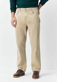 Man wearing beige straight-leg pants, dark brown leather shoes, and a dark green long-sleeve shirt with one hand in pocket.