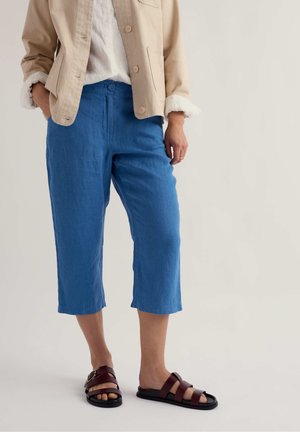 Persoană purtând o jachetă bej, cămașă albă, pantaloni scurți albaștri și sandale maro închis cu barete, stând pe un fundal neutră.