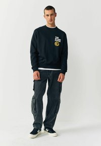 Marineblå sweatshirt med hvid tekst og rundt logo, parret med mørke cargo bukser med flere lommer og sorte sneakers.