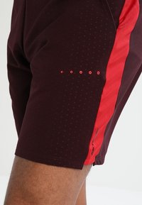 Nära bild av mörk vinröda sportshorts med små perforeringar och röd sidopanel, som bärs på en persons ben.