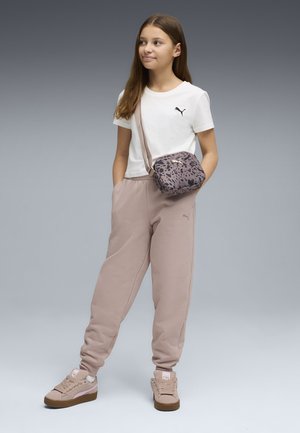 Ragazza giovane in piedi con le mani in tasca, indossa una maglietta bianca Puma, pantaloni beige, sneakers abbinate e una borsa a tracolla con stampa leopardata.