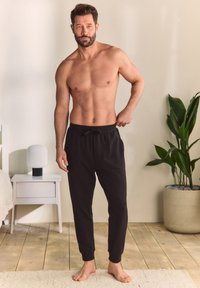 Pantaloni jogger negri, ușori, cu talie și mansete ajustabile cu șnur. Modelul stă desculț într-un interior luminos cu plante.
