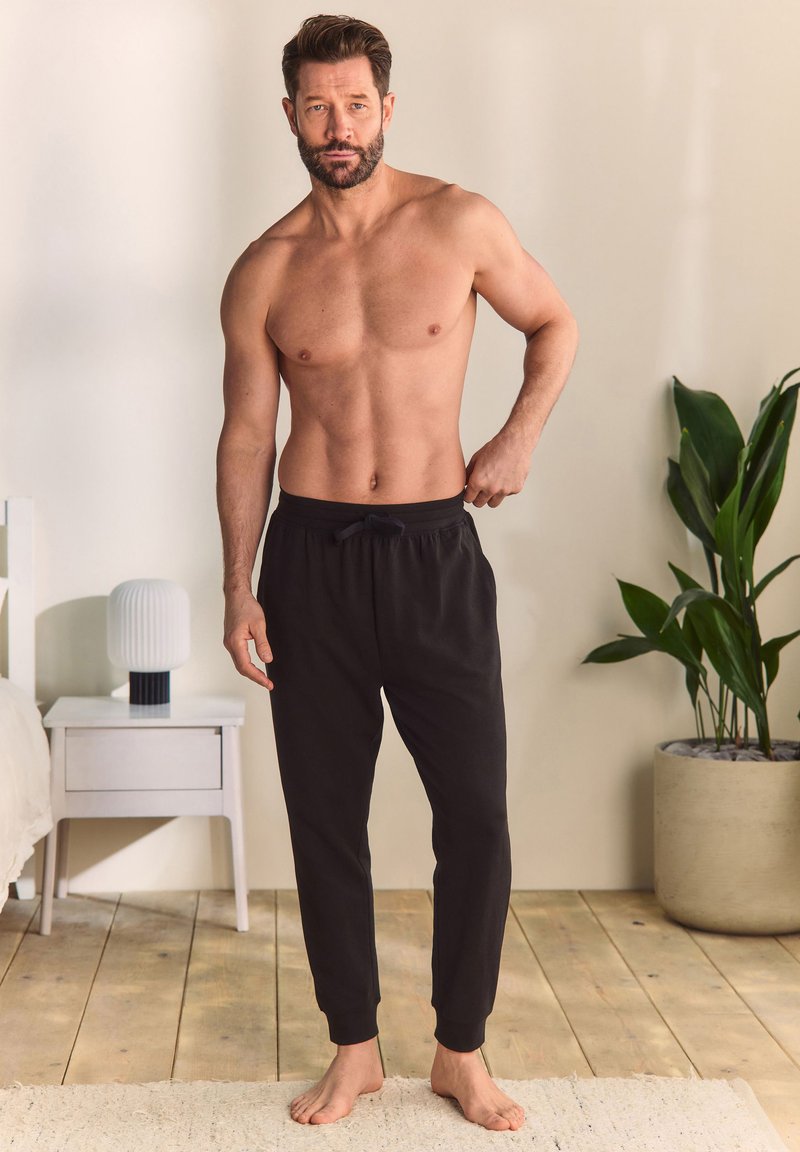 Black, lightweight jogger pants with adjustable drawstring waist and cuffs. Model stands barefoot in a bright interior with plants.