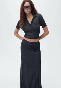 Haut polo cropped en tricot côtelé noir à manches courtes, associé à une jupe longue plissée assortie. Des lunettes de soleil mates complètent le look.