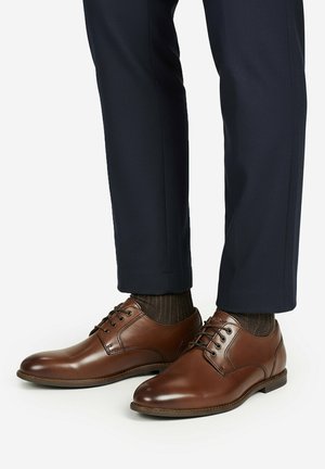 Braune Schnürschuhe aus Leder, getragen mit dunklen, gemusterten Socken und maßgeschneiderten marineblauen Hosen vor einem einfarbigen Hintergrund.