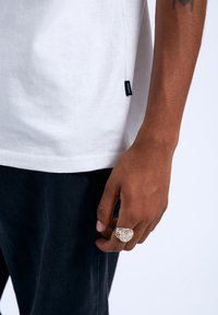 Bílý bavlněný tričko s božním štítkem, doplněné tmavými džíny. Na ruce se nachází texturovaný stříbrný prsten s propracovanými vzory.