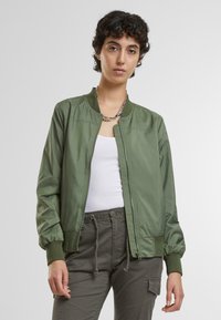 Olivovo zelená bomber bunda vyrobená z hladkého materiálu, s predným zipsom, rebrovanými manžetami a uvoľneným strihom. Model má na sebe biely top.