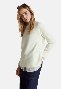 Sweter w kolorze kremowym, prążkowany, o luźnym kroju, z okrągłym dekoltem i długimi rękawami, noszony na paskowanym T-shircie.