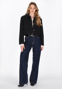 Schwarze, cropped Jacke mit zwei vorderen Taschen, kombiniert mit weit geschnittenen dunkelblauen Jeans mit Kontrastnähten und schwarzen Stiefeletten mit Absatz.