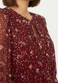 Blusa bordeaux con maniche trasparenti, motivo floreale in arancione, bianco e blu, con un dettaglio a fiocco sul collo e un fit rilassato.