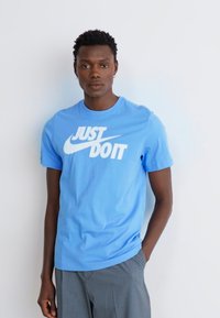 Camiseta de algodão azul clara com decote redondo, apresentando um grande logo branco "Just Do It" ao longo do peito. Mangas curtas e textura suave.
