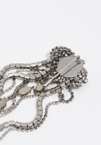 Metalowa broszka o okrągłej podstawie z wieloma segmentami łańcuszków. Zdobiona różnorodnymi błyszczącymi, prostokątnymi i kwadratowymi koralikami w srebrnych odcieniach.