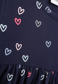 Marineblaues Kleid mit kurzen Ärmeln, das an der Unterseite gerafft ist. Verziert mit kleinen, mehrfarbigen Herzen in Rosa, Blau und Grau. Aus Baumwollstoff.