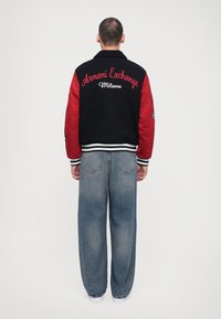Jachetă tip bomber neagră și roșie cu "Armani Exchange Milano" brodat pe spate, asortată cu blugi lejerați din denim albastru deschis.