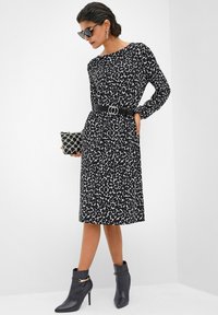 Frau in langärmligem Midi-Kleid mit schwarz-weißem Leopardenmuster, schwarzem Gürtel, schwarzen Stiefeletten, schwarzer Handtasche und Sonnenbrille, die posiert.