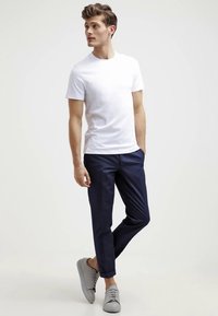 Fiatal férfi fehér, rövid ujjú pólóban, sötétkék felgöngyölt nadrágban és szürke sportcipőben áll, az egyik kezével a zsebében.