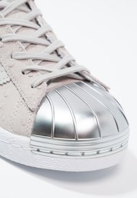 Sneaker in suede grigio chiaro con punta metallica argento, superiore texturizzato e lacci grigi. Presenta cuciture distintive e suola in gomma.