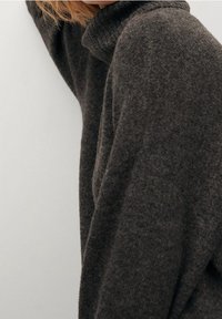 Un pull col roulé gris foncé en tissu doux et texturé avec une coupe décontractée. Il présente des épaules tombantes et des détails côtelés au niveau du col.