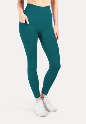 Leggings taille haute teal avec une poche latérale, fabriqués en tissu extensible au toucher doux. Accompagnés de chaussures de sport blanches.