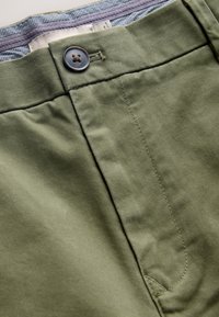 Gros plan d'un pantalon vert olive avec une ceinture boutonnée et une doublure intérieure à carreaux visible sur le bord supérieur.