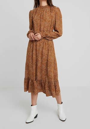 Kvinde iført en langærmet, ankellang brun kjole med hvid blomstermønster og hvide ankelstøvler, stående foran en ensfarvet grå baggrund.