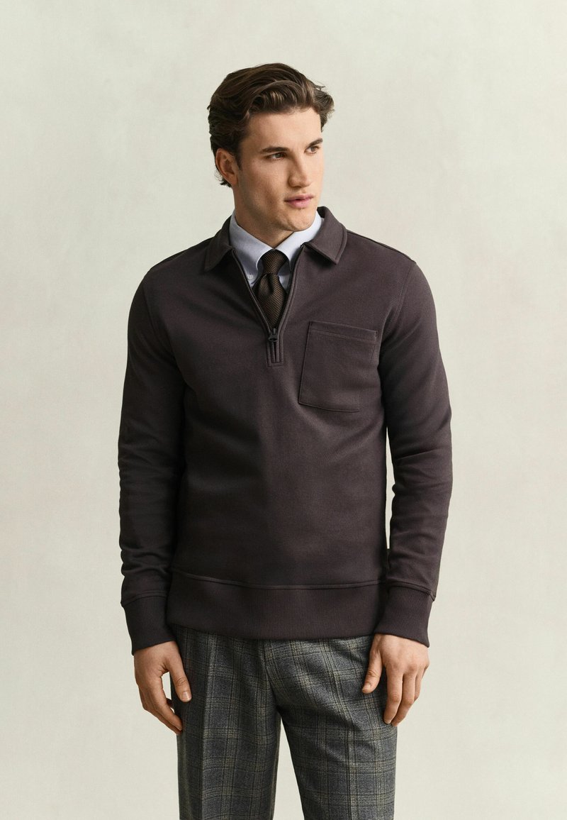 Homme portant un pull marron foncé à fermeture quart de zip sur une chemise bleue et une cravate foncée, associé à un pantalon gris à carreaux, debout devant un fond uni et clair.