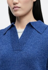 Blauer Strickpullover mit V-Ausschnitt und strukturiertem Kragen, über einem hellgrauen Oberteil getragen. Weiche Textur und gleichmäßige Nahtdetails sind sichtbar.