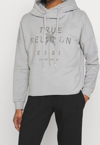 Felpa grigia corta con cappuccio oversize, lacci e stampa metallica con la scritta "TRUE RELIGION 2002" e dettaglio di palma.