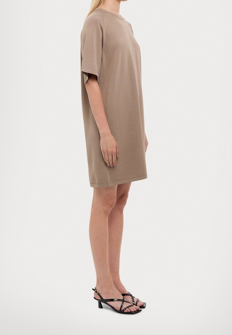 Femme portant une robe beige ample jusqu’aux genoux et des sandales noires à talons bas et à brides, debout de profil sur un fond blanc.