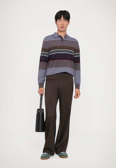 Brun og lilla stribet langærmet polo shirt, brune bukser, grønne og grå sko, holder en sort cylindrisk taske.
