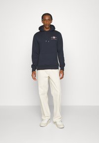 Marinblå hoodie med dragsko och framficka, med ett logotryck. Matchas med krämfärgade vida byxor och vita sneakers.