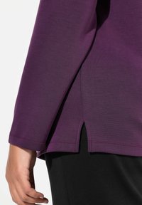 Camicia a maniche lunghe in tessuto viola ricco con una texture liscia, caratterizzata da uno spacco laterale e cuciture a contrasto. Tessuto nero visibile all'orlo.