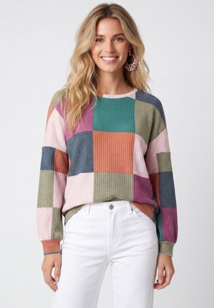 Lächelnde Frau mit langen blonden Haaren, die einen bunten Patchwork-Pullover und weiße Hose trägt, stehend vor einem schlichten, hellen Hintergrund.