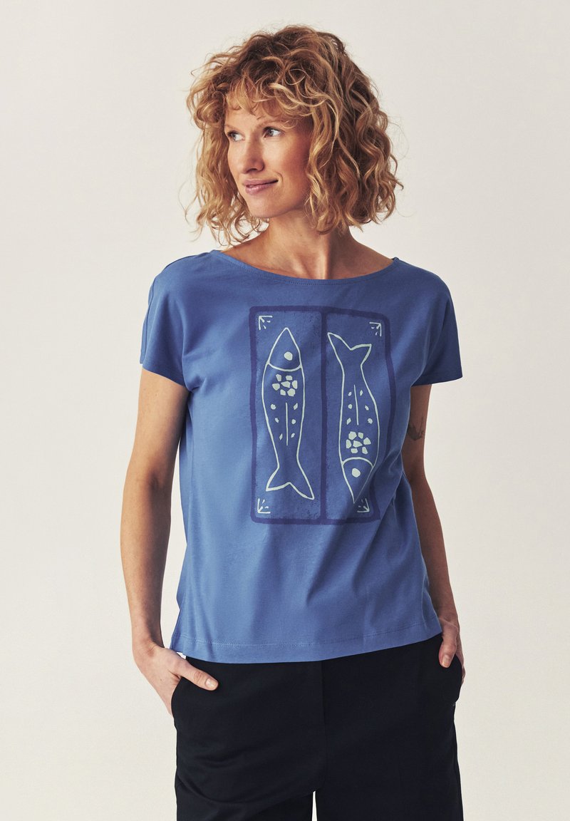Femme aux cheveux bouclés portant un T-shirt bleu avec deux imprimés stylisés de poissons, mains dans les poches, regardant de côté sur un fond uni.