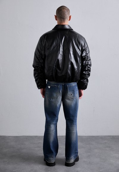 Homme avec des cheveux très courts portant une veste noire brillante et un jean bleu délavé, faisant face à un mur gris clair uni sur un sol gris.