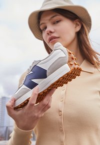 Ung kvinna som bär en beige hatt och ribbade kofta håller upp en vit och grå sneaker med blå logotyp och orange texturerad sula.
