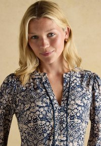 Lillelille sinine ja kreemjas lillemustriga pluus, millel on puffsuletud varrukad, V-kaelus ja sinine ääris nööpide juures. Pehme kangas botaanilise prindiga.