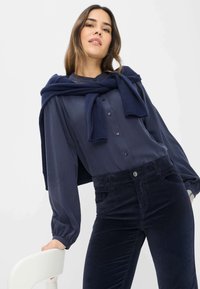 Blouse bleu marine avec des fermetures à boutons et des manches ballon, associée à un pantalon en velours côtelé bleu marine foncé. Pull drapé de manière décontractée autour des épaules.