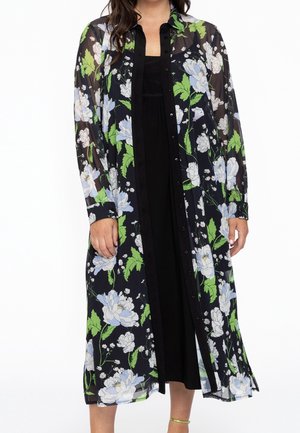 Vrouw draagt een lange, doorschijnende zwarte jurk met knopen, bedrukt met groene bladeren en witte bloemen, over een effen zwarte jurk.