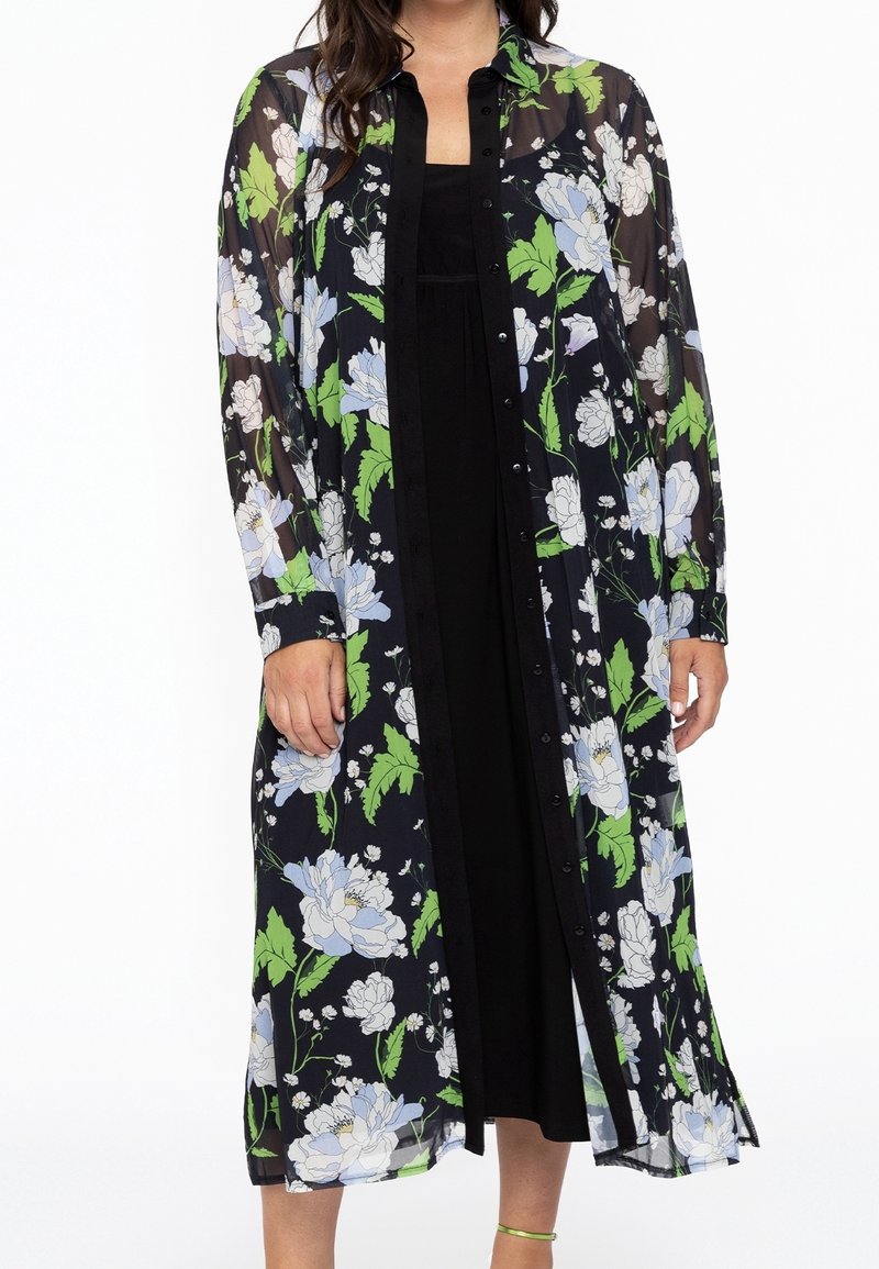 Femme portant une longue robe noire transparente boutonnée ornée de feuilles vertes et de fleurs blanches, portée sur une robe noire unie.