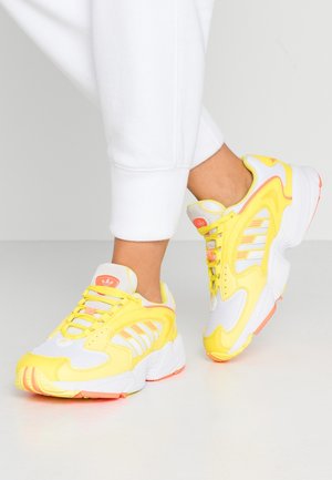 Person iført gule og hvide Adidas-sneakers med orange accenter og hvide bukser, stående på en lysegrå gulv.