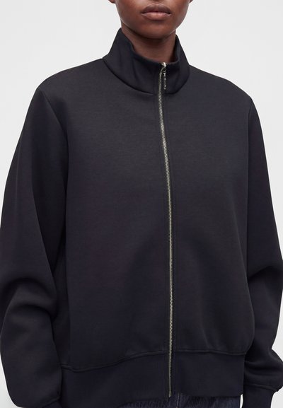 Personne portant une veste noire zippée avec un col montant et des poignets et un ourlet côtelés, montrée de la poitrine au cou.